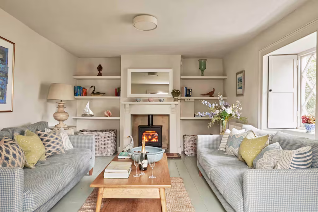 The cosy living room interior of Cove Cottage at Lulworth Cove in Dorset. A wood buner contains a roaring fire, backed by alcove shelves. Two pale blue sofas sit in the foreground around a wooden coffee table The room is bright and airy with warm, magnolia tones on the walls