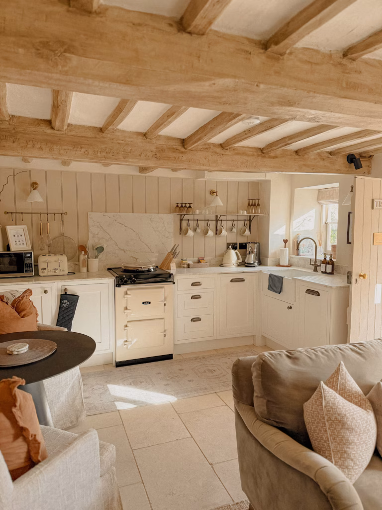 The open-plan kitchen at Tregoney Cottage in the Cotswolds, with cream shaker cabinets, a cream Aga, marble splashback, brass hanging rail with white mugs, exposed oak beams overhead, and stone tile flooring, with a round dining table and linen sofas in the foreground
