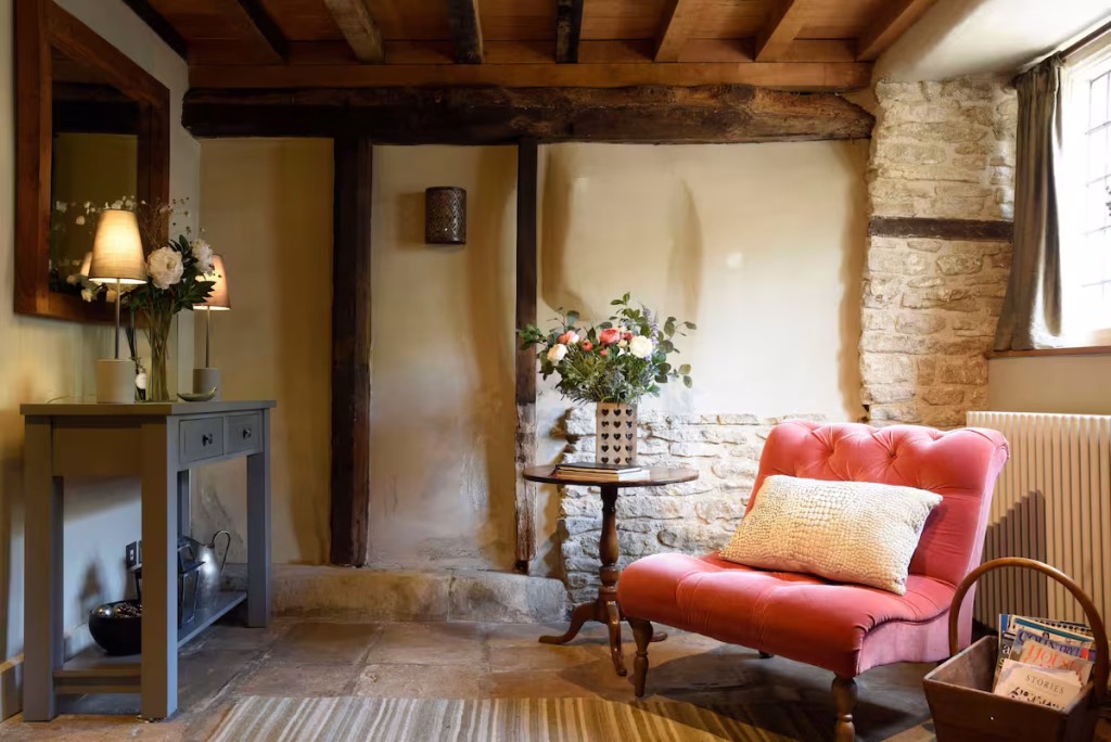 A cosy sitting room corner at The Gates in Castle Combe, with ancient flagstone floors, exposed Cotswold stone walls, heavy oak ceiling beams, a coral pink tufted armchair with a textured cushion, a pedestal side table with a fresh flower arrangement, and a grey painted console table with a lamp reflected in a large mirror