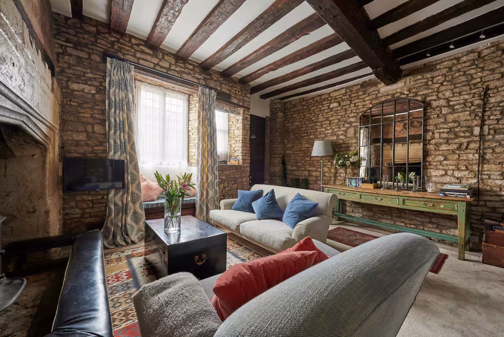 The living room at The Old Court House in Castle Combe, with floor-to-ceiling exposed Cotswold stone walls, heavy dark oak ceiling beams, a cream sofa with blue cushions, a grey sofa with red cushions, a black metal trunk as a coffee table, an antique green painted console table, and patterned curtains framing a leaded window