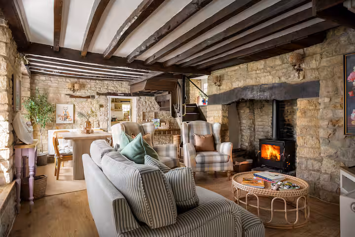 The living room of Jasmine Cottage in the Cotswolds, with exposed Cotswold stone walls, heavy wooden ceiling beams, a lit wood-burning stove set into an inglenook fireplace, a striped sofa and plaid armchairs