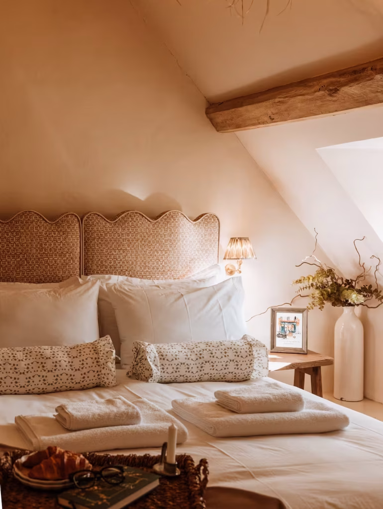 A sun-warmed bedroom at Tregoney Cottage in the Cotswolds, with a scallop-edged printed headboard, white linen, floral bolster cushions, a brass wall light, woven rattan light fitting overhead, exposed beam, and a large ceramic vase with dried foliage on a rustic bedside stool