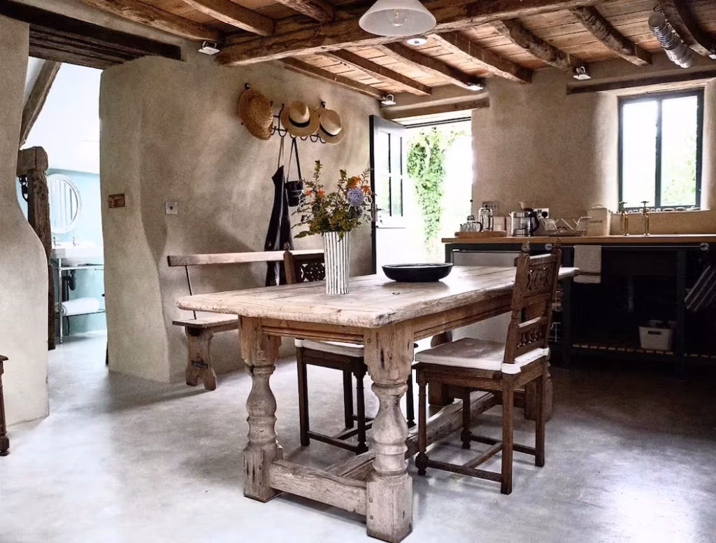 The kitchen and dining area of The Barn on Dartmoor, with rough lime-plastered walls, heavy wooden ceiling beams, a large bleached wood refectory table, antique chairs and a dark navy kitchen beyond French doors opening to the garden