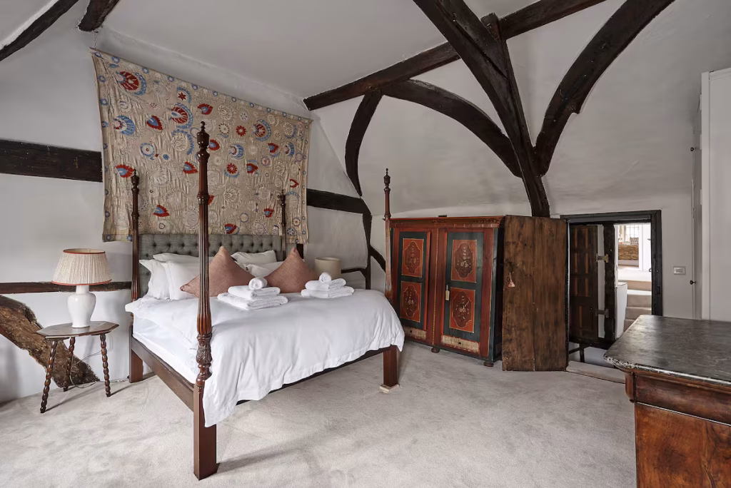 A characterful bedroom at The Old Court House in Castle Combe, with a dark mahogany four-poster bed, white linen, dusty pink cushions, a large antique painted wardrobe in red and black, a dramatic arched framework of heavy oak beams overhead, and an embroidered textile hanging as a canopy behind the bed