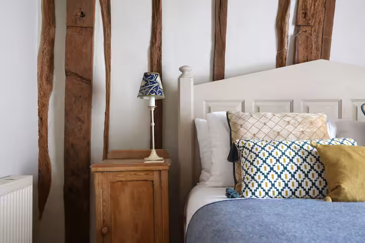 A bedroom in a medieval cottage in Sandwich, Kent, with characterful twisted oak beams against white walls, a white painted headboard, mustard and teal patterned cushions and a William Morris lampshade on the bedside table
