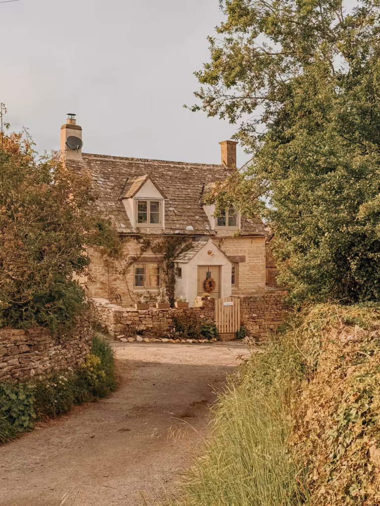 The exterior of Tregoney Cottage in the Cotswolds at golden hour, a honey-coloured stone cottage with a steep slate roof, dormer windows, climbing plants around the front door, a wreath on the gate, and terracotta pots, framed by mature trees and a dry stone wall along a country lane