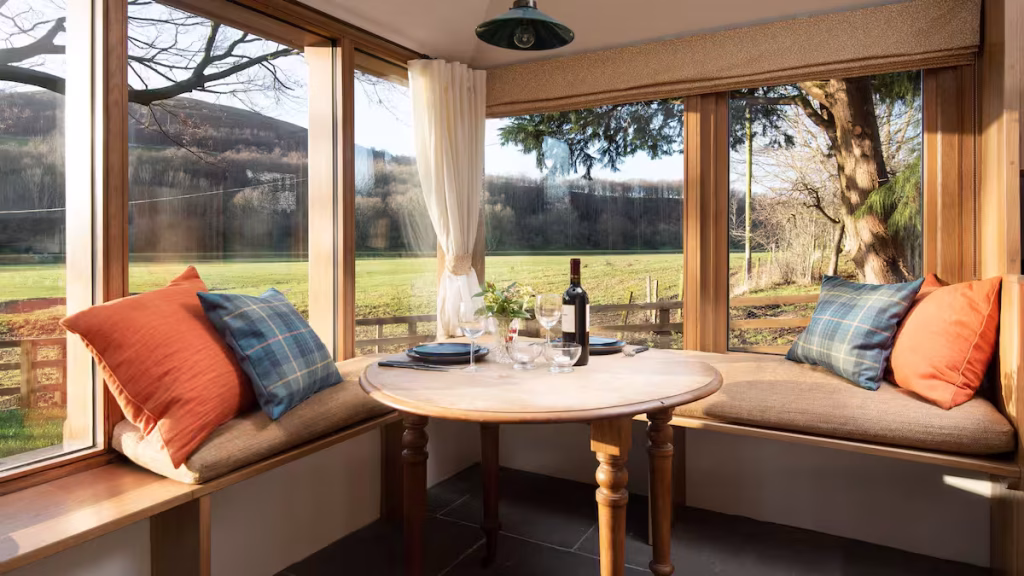 dining-area-dovecote-reedsford.jpg A wrap-around window seat dining nook at The Dovecote at Reedsford, with orange and tartan cushions, a round wooden table set with wine glasses and plates, and wide views across a green field and trees in winter sun