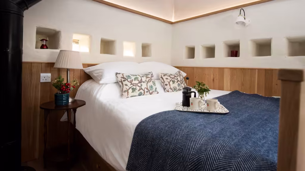 The bedroom at The Dovecote at Reedsford, with white plastered walls, a row of illuminated square alcoves above oak panelling, a double bed with navy herringbone throw and floral cushions, and a French press coffee tray