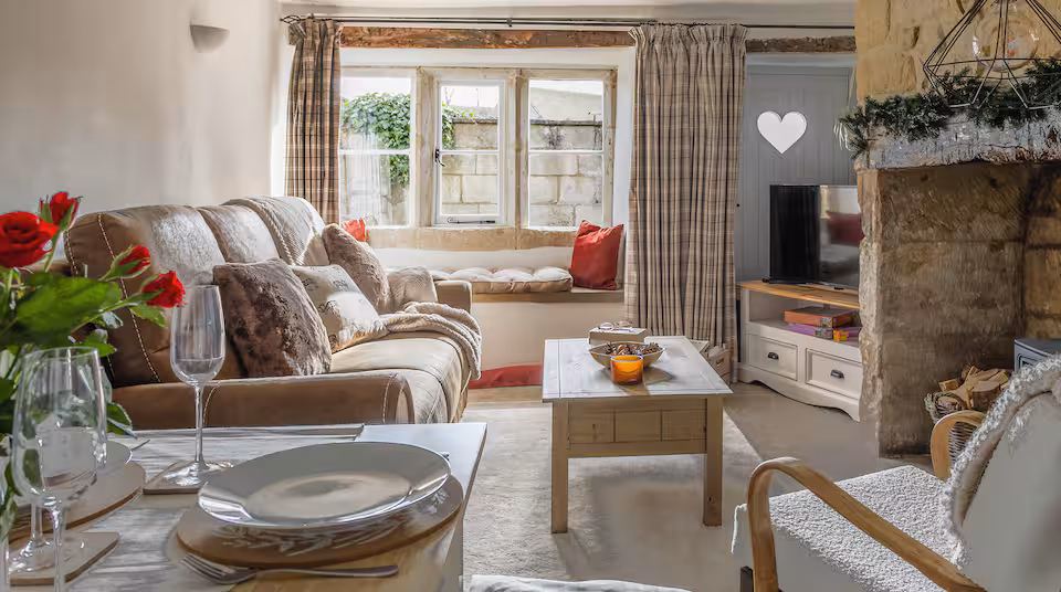 The living room at Love View Cottage in Painswick, with a stone fireplace, tartan curtains, a cushioned window seat, cream sofa and a dining table set with glasses and plates in the foreground